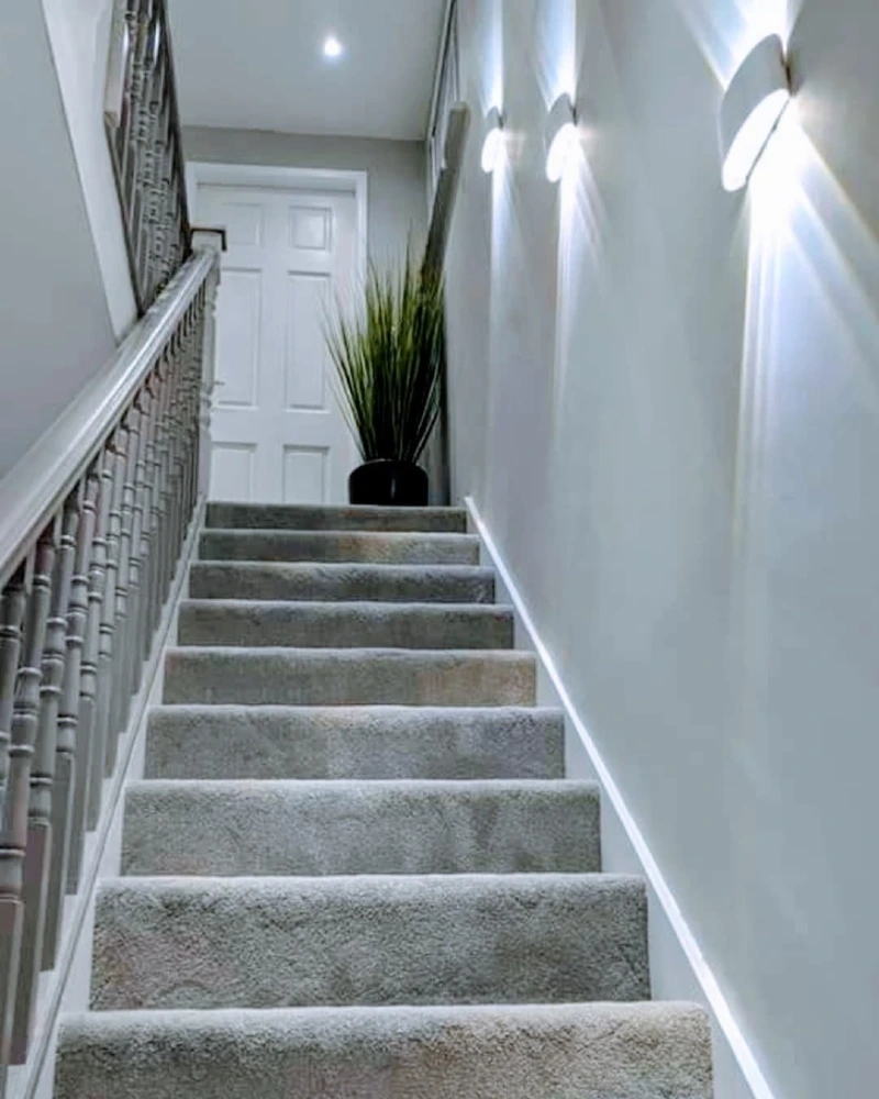 Modern White Sconces Illumining a Grey Carpeted Staircase and Top Landing Planter
