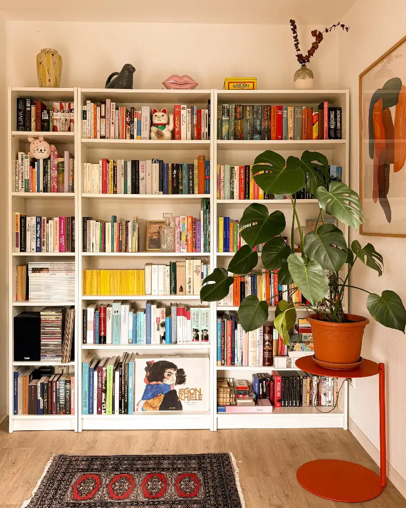 White Book Wall with Monstera Leaves, Red Side Table, and Playful Shelf Objects