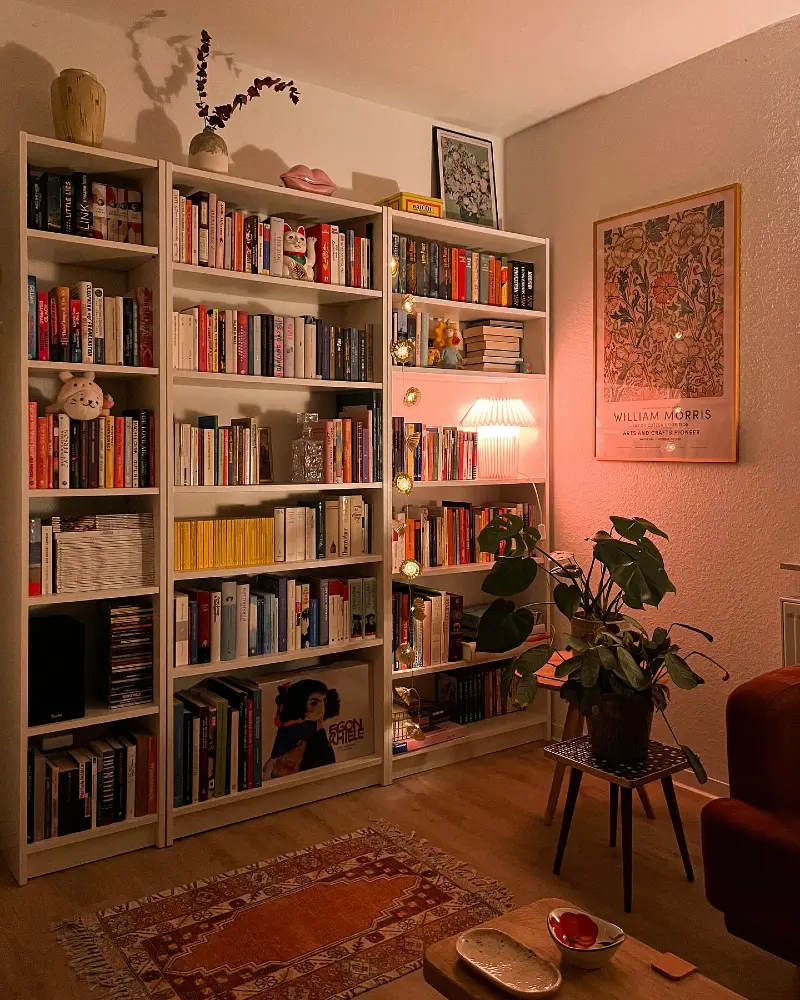 White Bookcase Wall with Amber Lamp Glow and William Morris Art